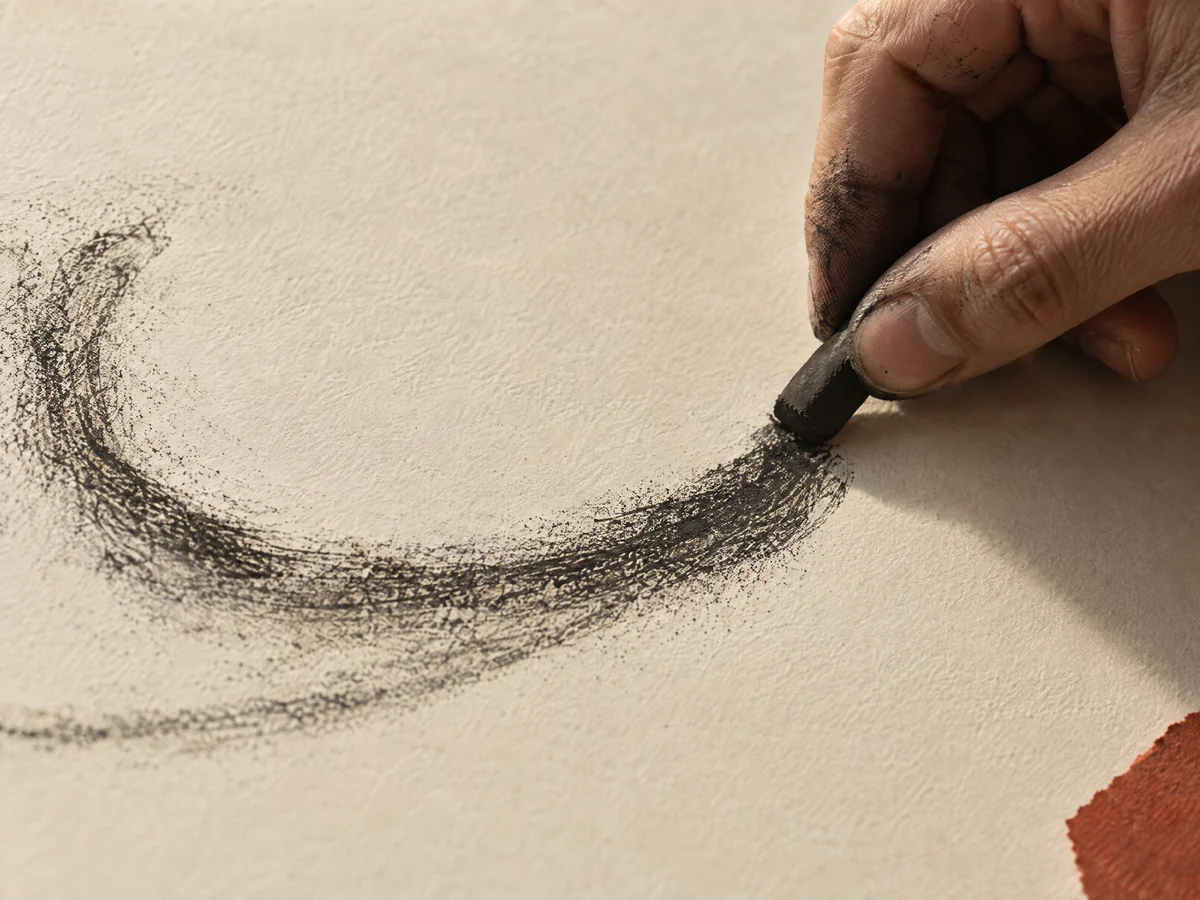 A charcoal-dusted hand drawing an expressive curved mark on ivory paper.
